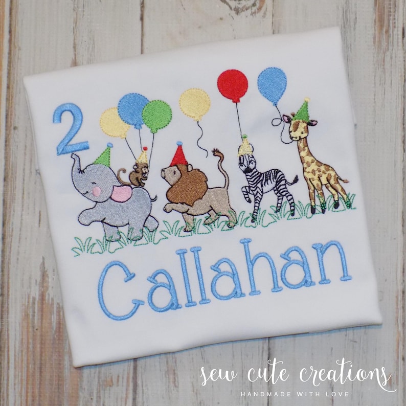 Puede incluir: Una camiseta blanca con un dise&ntilde;o bordado de un elefante gris, un le&oacute;n marr&oacute;n, una cebra blanca y negra y una jirafa beige, todos con gorros de fiesta. El dise&ntilde;o incluye globos de colores, el n&uacute;mero 2 y el nombre Callahan en azul.