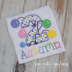 May include: White t-shirt with a colorful embroidered design for a second birthday. The number "2" is decorated with polka dots and surrounded by bubbles. The name "Autumn" is embroidered in rainbow colors.