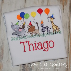 Puede incluir: Una camiseta blanca con un dise&ntilde;o bordado de un elefante gris, un mono marr&oacute;n, un le&oacute;n beige, una cebra blanca y negra y una jirafa amarilla y marr&oacute;n, todos con gorros de fiesta y globos de colores sobre el nombre rojo Thiago.