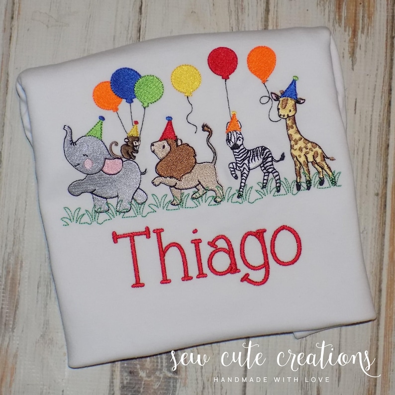 Puede incluir: Una camiseta blanca con un dise&ntilde;o bordado de un elefante gris, un mono marr&oacute;n, un le&oacute;n beige, una cebra blanca y negra y una jirafa amarilla y marr&oacute;n, todos con gorros de fiesta y globos de colores sobre el nombre rojo Thiago.