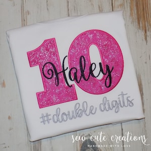 Puede incluir: Una camiseta blanca con una gran aplicación de purpurina rosa en forma de número 10. El nombre Haley está bordado en cursiva negra sobre el centro del número. Debajo del número, el texto #double digits está bordado en hilo plateado.