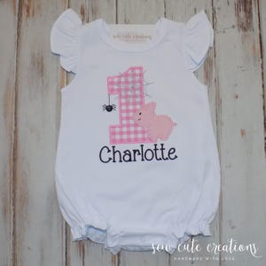 May include: White baby romper with ruffled sleeves. Features a pink gingham number 1, a pink pig, and the name "Charlotte" in black script. A small spider and spiderweb are also included in the design. The text "sew cute creations" is printed on the top.