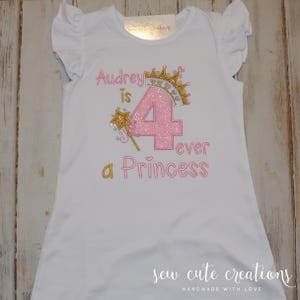 May include: White dress with ruffled sleeves, featuring the text "Audrey is 4 ever a Princess" in pink and gold embroidery. Includes a crown and wand design. "Sew Cute Creations" is printed at the bottom.