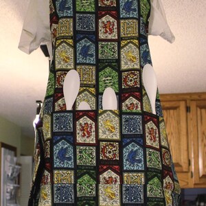 May include: A colorful apron with a repeating pattern of crests in red, blue, green, and yellow. The apron has a black neck strap and pockets, with white spoons in the pockets. The apron is displayed on a mannequin.