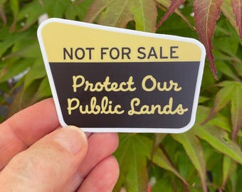May include: A sticker with a yellow and black design, featuring the text "NOT FOR SALE" above "Protect Our Public Lands" in a handwritten style. The sticker is held against a backdrop of green and red leaves.