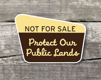 May include: A sticker with a tan top section and a brown bottom section. The text "NOT FOR SALE" is in the tan section. The brown section has the text "Protect Our Public Lands" in a cursive font. The sticker is on a wooden surface.
