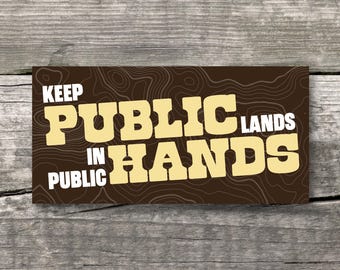 May include: A rectangular sticker with a dark brown background featuring a thin, light brown topographic map line pattern. The text reads KEEP PUBLIC LANDS IN PUBLIC HANDS in bold, light yellow, block-style lettering across the center.