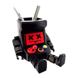 May include: A black desk organizer shaped like a retro arcade cabinet with articulated arms and legs. The screen features a red display with two black X symbols for eyes and a straight line for a mouth. It includes a red joystick and two buttons.