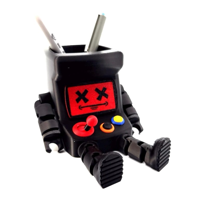 May include: A black desk organizer shaped like a retro arcade cabinet with articulated arms and legs. The screen features a red display with two black X symbols for eyes and a straight line for a mouth. It includes a red joystick and two buttons.