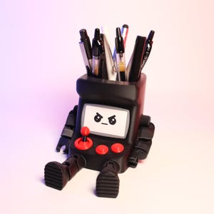 May include: A black desk organizer shaped like a retro arcade cabinet with articulated arms and legs. The front features a screen with an angry face graphic, a red joystick, and two red buttons. Several pens and markers are stored in the top opening.