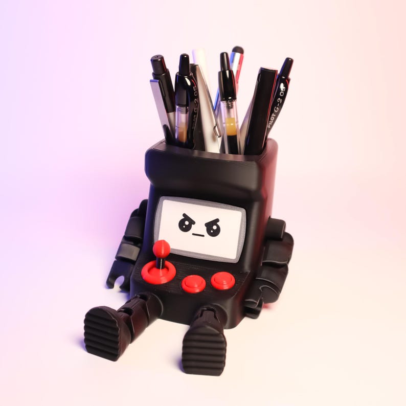 May include: A black desk organizer shaped like a retro arcade cabinet with articulated arms and legs. The front features a screen with an angry face graphic, a red joystick, and two red buttons. Several pens and markers are stored in the top opening.