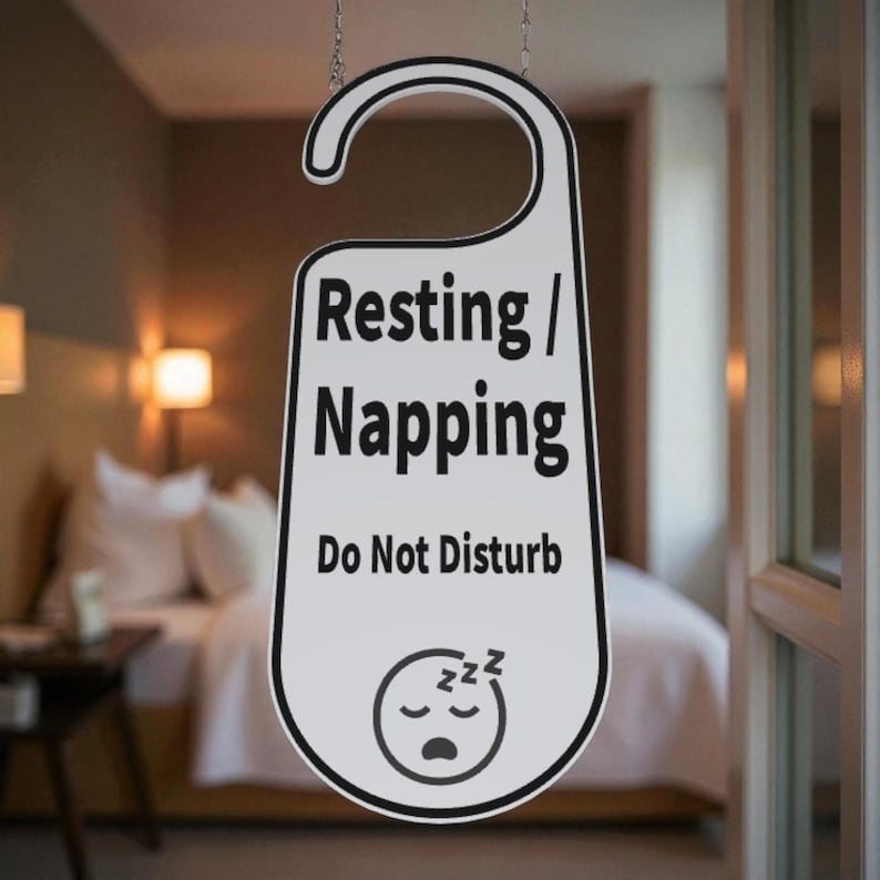 K&ouml;nnte beinhalten: Wei&szlig;er und schwarzer T&uuml;ranh&auml;nger mit der Aufschrift "Resting / Napping Do Not Disturb". Der Anh&auml;nger hat eine gebogene Oberseite zum Aufh&auml;ngen und eine Grafik eines schlafenden Gesichts. Im Hintergrund ist ein Hotelzimmer zu sehen.