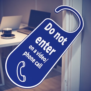 K&ouml;nnte beinhalten: Ein blauer T&uuml;rh&auml;nger mit wei&szlig;em Text "Do not enter on a video/phone call". Unten befindet sich ein wei&szlig;es Telefonsymbol. Im Hintergrund ist ein Schreibtisch mit Laptop und Kaffeetasse zu sehen.