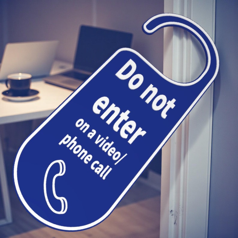 K&ouml;nnte beinhalten: Ein blauer T&uuml;rh&auml;nger mit wei&szlig;em Text "Do not enter on a video/phone call". Unten befindet sich ein wei&szlig;es Telefonsymbol. Im Hintergrund ist ein Schreibtisch mit Laptop und Kaffeetasse zu sehen.