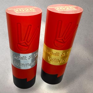Peut inclure: Deux trophées de badminton rouges avec des bases noires. Les trophées présentent un symbole de main et le texte "WELLINGBOROUGH 2025 BADMINTON". L'un des trophées indique "Adult & Runner Up" et l'autre "Adult & Winner".