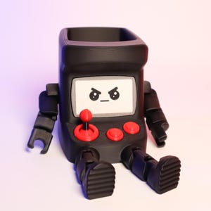 May include: A matte black desk organizer shaped like a retro arcade cabinet with articulated arms and legs. The screen features a printed angry face, and the control panel includes a red joystick and two red buttons. The top is open for storage.