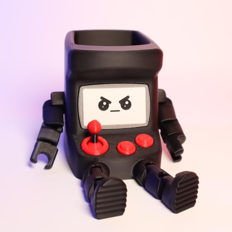 May include: A matte black desk organizer shaped like a retro arcade cabinet with articulated arms and legs. The screen features a printed angry face, and the control panel includes a red joystick and two red buttons. The top is open for storage.