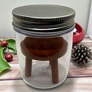 May include: A clear glass jar with a dark metal lid. Inside the jar is a small, brown, three-legged stool. The jar is sitting on a wooden surface, with red berries and a pinecone in the background.