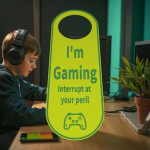 K&ouml;nnte beinhalten: Ein hellgr&uuml;ner T&uuml;rh&auml;nger mit dem Text "I'm Gaming, Interrupt at your peril" und einem Gamecontroller-Symbol. Im Hintergrund ist eine Person mit Kopfh&ouml;rern zu sehen, die sich auf einen Bildschirm konzentriert.
