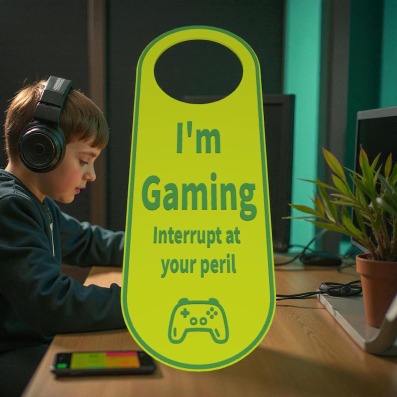 K&ouml;nnte beinhalten: Ein hellgr&uuml;ner T&uuml;rh&auml;nger mit dem Text "I'm Gaming, Interrupt at your peril" und einem Gamecontroller-Symbol. Im Hintergrund ist eine Person mit Kopfh&ouml;rern zu sehen, die sich auf einen Bildschirm konzentriert.