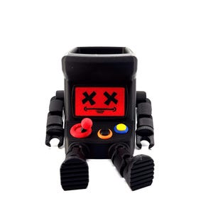 May include: A black robot-shaped desk organizer featuring a red screen with two black X-shaped eyes and a curved mouth. The front includes a red joystick, a black button with an orange ring, and a yellow button. The robot has articulated arms and feet.