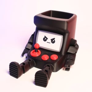 May include: A black, hollow, arcade-themed desk organizer shaped like a seated robot. It features a white screen with an angry face graphic, a red joystick, and two red buttons on the front. The robot has articulated black arms and feet.
