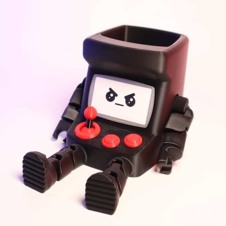 May include: A black, hollow, arcade-themed desk organizer shaped like a seated robot. It features a white screen with an angry face graphic, a red joystick, and two red buttons on the front. The robot has articulated black arms and feet.