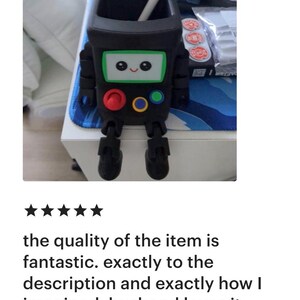 May include: A black, square-shaped desk organizer designed to resemble a small robot with articulated legs. The front features a white screen with a smiling face, a red button, a yellow button, and a blue button. A white stylus rests inside the top.