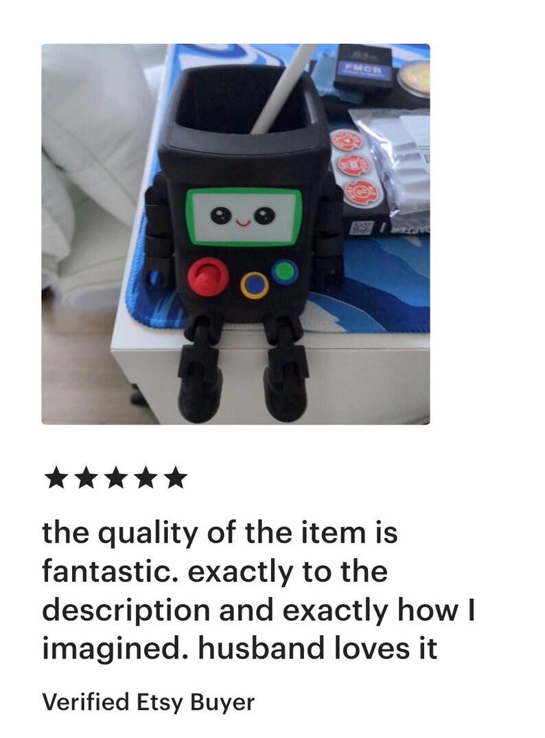 May include: A black, square-shaped desk organizer designed to resemble a small robot with articulated legs. The front features a white screen with a smiling face, a red button, a yellow button, and a blue button. A white stylus rests inside the top.