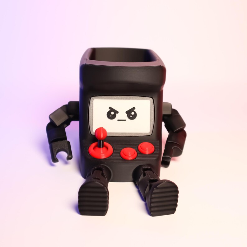 May include: A matte black arcade cabinet figurine with articulated arms and legs. The screen features a printed angry face with furrowed brows. The control panel includes a red joystick and two red buttons. The object has a hollow top opening.