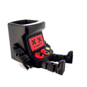 May include: A black plastic desk organizer shaped like a seated robot with an arcade cabinet head. The robot features a red screen with black X eyes and a smiling mouth, a red joystick, and blue and orange circular buttons on its chest area.