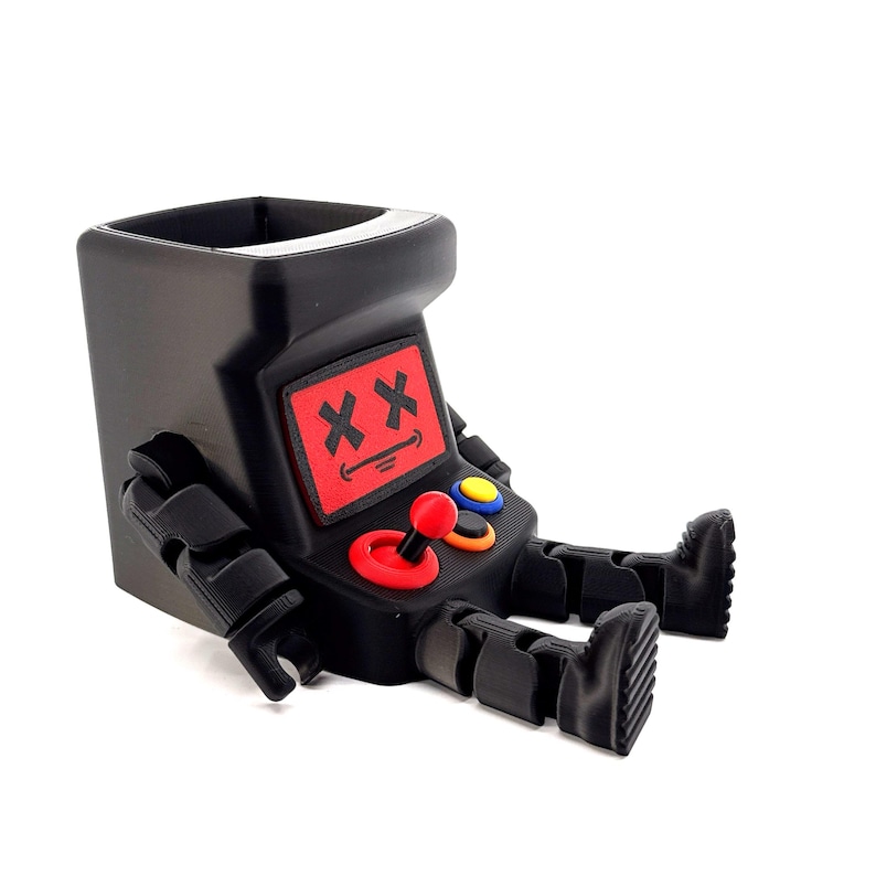 May include: A black plastic desk organizer shaped like a seated robot with an arcade cabinet head. The robot features a red screen with black X eyes and a smiling mouth, a red joystick, and blue and orange circular buttons on its chest area.