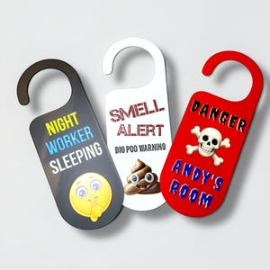 May include: Three novelty door hangers. One is black with "NIGHT WORKER SLEEPING" text and a yellow emoji. Another is white with "SMELL ALERT" text and a poop emoji. The third is red with "DANGER ANDY'S ROOM" text and a skull.