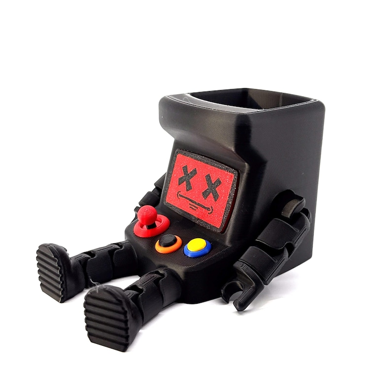 May include: A black desk organizer shaped like a seated arcade cabinet with articulated arms and legs. The screen features a red panel with black X eyes and a smiling mouth. The control panel includes a red joystick, a black button, and a yellow button.
