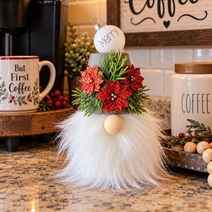 May include: A decorative gnome figure featuring a gray pot hat topped with a white ornament labeled Merry, adorned with red glittery poinsettia flowers and green pine sprigs, and a long white faux fur beard with a round wooden nose.