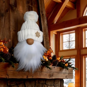 May include: A white gnome decoration with a knitted hat, a fluffy white beard, and a wooden nose. The hat features a gold snowflake design. The gnome is displayed on a wooden mantelpiece with festive greenery and pinecones.