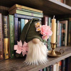 May include: A decorative gnome with a green hat, white beard, and wooden nose. The gnome is adorned with pink flowers and a pink bow. A lit candle in a brass holder and a collection of books are in the background.
