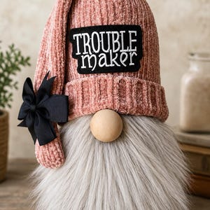May include: A decorative gnome figurine featuring a dusty pink ribbed velvet hat with a black bow on the side. A black rectangular patch with the white embroidered text TROUBLE maker is centered on the hat. It has a round wooden nose and a long gray faux fur beard.