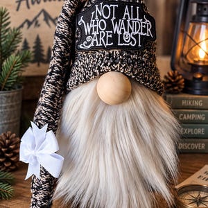 May include: A gnome figurine with a long white faux fur beard and a wooden bead nose. It wears a tall, slouchy black and tan knit hat featuring a black patch with the white embroidered text "NOT ALL WHO WANDER ARE LOST" and a white ribbon bow.