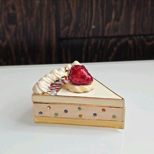May include: A decorative trinket box shaped like a slice of cake. The box features a cream-colored top with a red strawberry on top. The base is a light beige with colorful gems and gold trim.