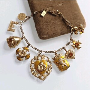 May include: A gold-toned charm bracelet with various detailed charms. Charms include a bell, a letter "E", a heart, a crown with a lion, and a fleur-de-lis. The bracelet is on a brown surface.