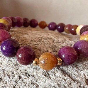 May include: A beaded bracelet featuring polished spherical beads in shades of deep purple, magenta, and golden yellow. The design includes small, shiny gold-toned metal spacers placed between the beads, creating a repeating pattern of colors.