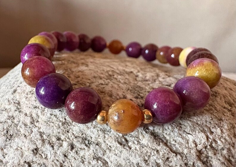 May include: A beaded bracelet featuring polished spherical beads in shades of deep purple, magenta, and golden yellow. The design includes small, shiny gold-toned metal spacers placed between the beads, creating a repeating pattern of colors.