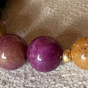 May include: A beaded bracelet featuring smooth, spherical beads in shades of deep purple, mustard yellow, and brownish-gold. The beads are arranged in a repeating pattern and are separated by small, polished gold-toned metal spacer beads.