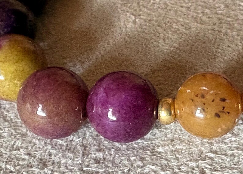 May include: A beaded bracelet featuring smooth, spherical beads in shades of deep purple, mustard yellow, and brownish-gold. The beads are arranged in a repeating pattern and are separated by small, polished gold-toned metal spacer beads.