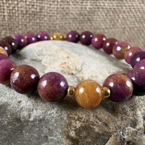 May include: A beaded bracelet featuring smooth, round purple beads and a single, larger, polished golden-yellow bead at the center. The beads are separated by small, shiny gold-toned metal spacers, creating a consistent, circular pattern throughout the piece.