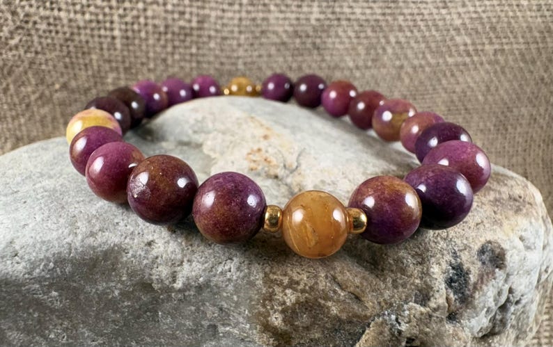 May include: A beaded bracelet featuring smooth, round purple beads and a single, larger, polished golden-yellow bead at the center. The beads are separated by small, shiny gold-toned metal spacers, creating a consistent, circular pattern throughout the piece.