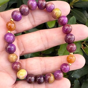 May include: A beaded bracelet featuring round, polished stones in shades of deep purple, magenta, and golden yellow. The bracelet is accented with small, smooth, metallic gold-toned spherical spacers placed between the colorful gemstone beads.