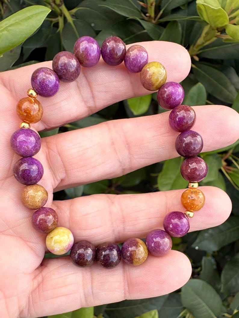 May include: A beaded bracelet featuring round, polished stones in shades of deep purple, magenta, and golden yellow. The bracelet is accented with small, smooth, metallic gold-toned spherical spacers placed between the colorful gemstone beads.