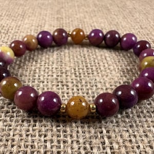 May include: A beaded bracelet featuring smooth, round beads in shades of deep purple, magenta, and mustard yellow. The beads are arranged in a circular pattern and are accented by small, polished gold-toned metal spacers placed between the beads.